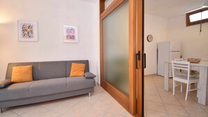 A modern living room with a grey sofa, two cushions, a wooden-framed sliding door, a white refrigerator, a dining table with chairs, and two framed artworks on the wall. - Three-room apartment downtown with two bathrooms and A/C (Santa Teresa Gallura)