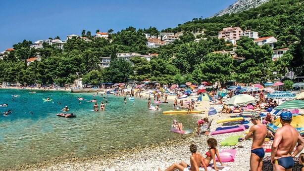 A crowded beach with clear turquoise water, people swimming and sunbathing, and buildings in the background.