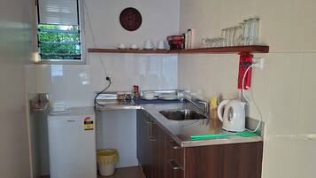 Une cuisine de taille réduite, équipée d’un réfrigérateur, d’un évier et d’une fenêtre avec des stores.