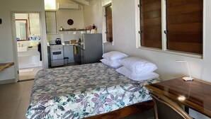 Ein Schlafzimmer mit Bett, Schreibtisch, einem Fenster mit Holzjalousien und einer kleinen Küchenzeile im Hintergrund.