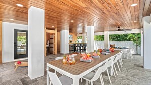 Un vaste espace extérieur aménagé pour les repas, avec une longue table en bois, des chaises blanches et un ventilateur de plafond.