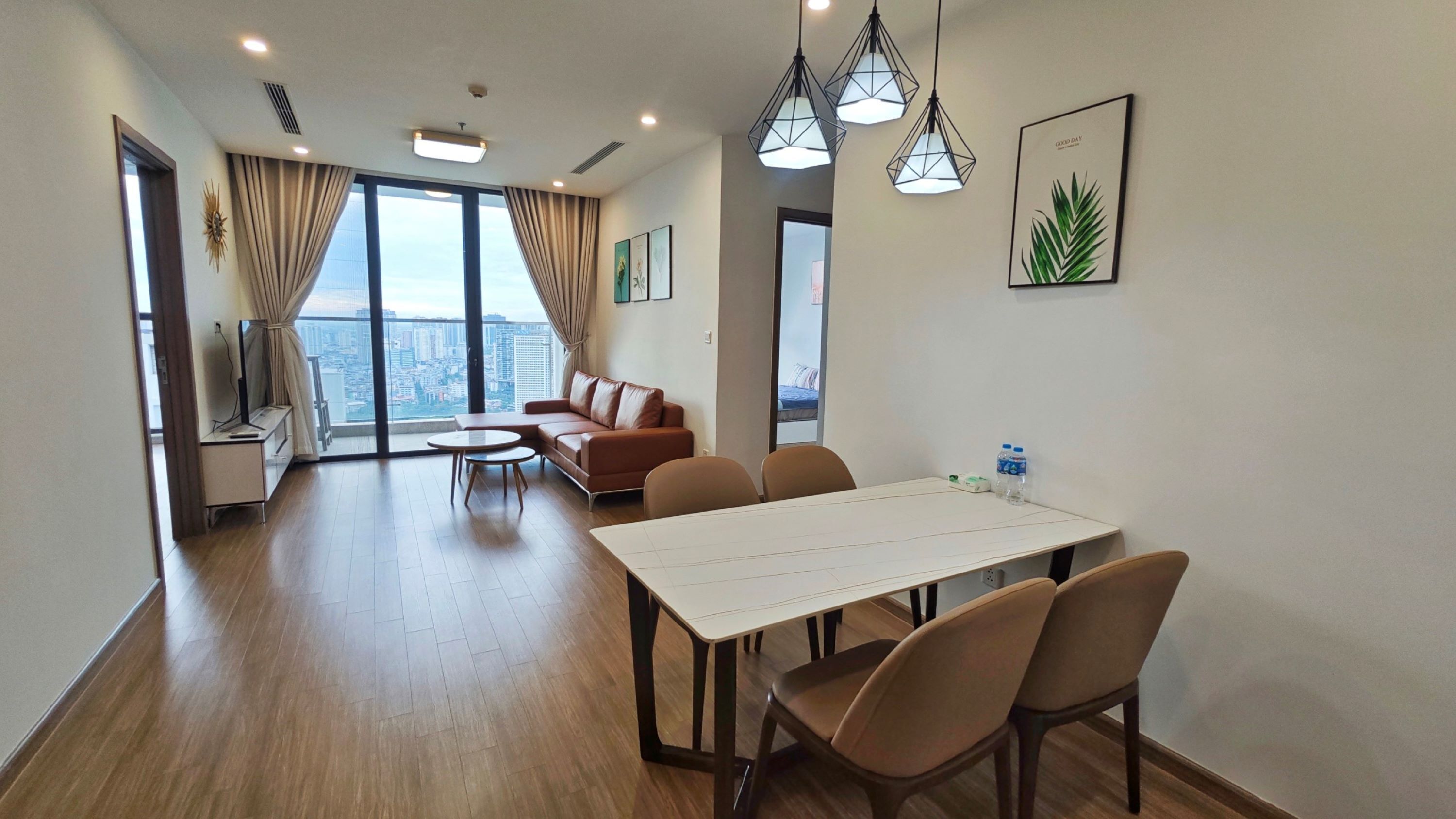 Habitación de hotel moderna con un ventanal amplio que ofrece vistas a la ciudad, un sofá, una mesa de comedor con sillas y un televisor sobre un mueble.