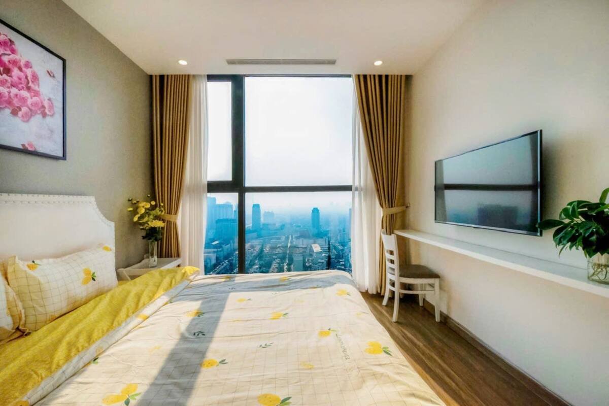 A bedroom with a large window, a bed with a floral bedspread, a TV on the wall, and a small desk with a chair.