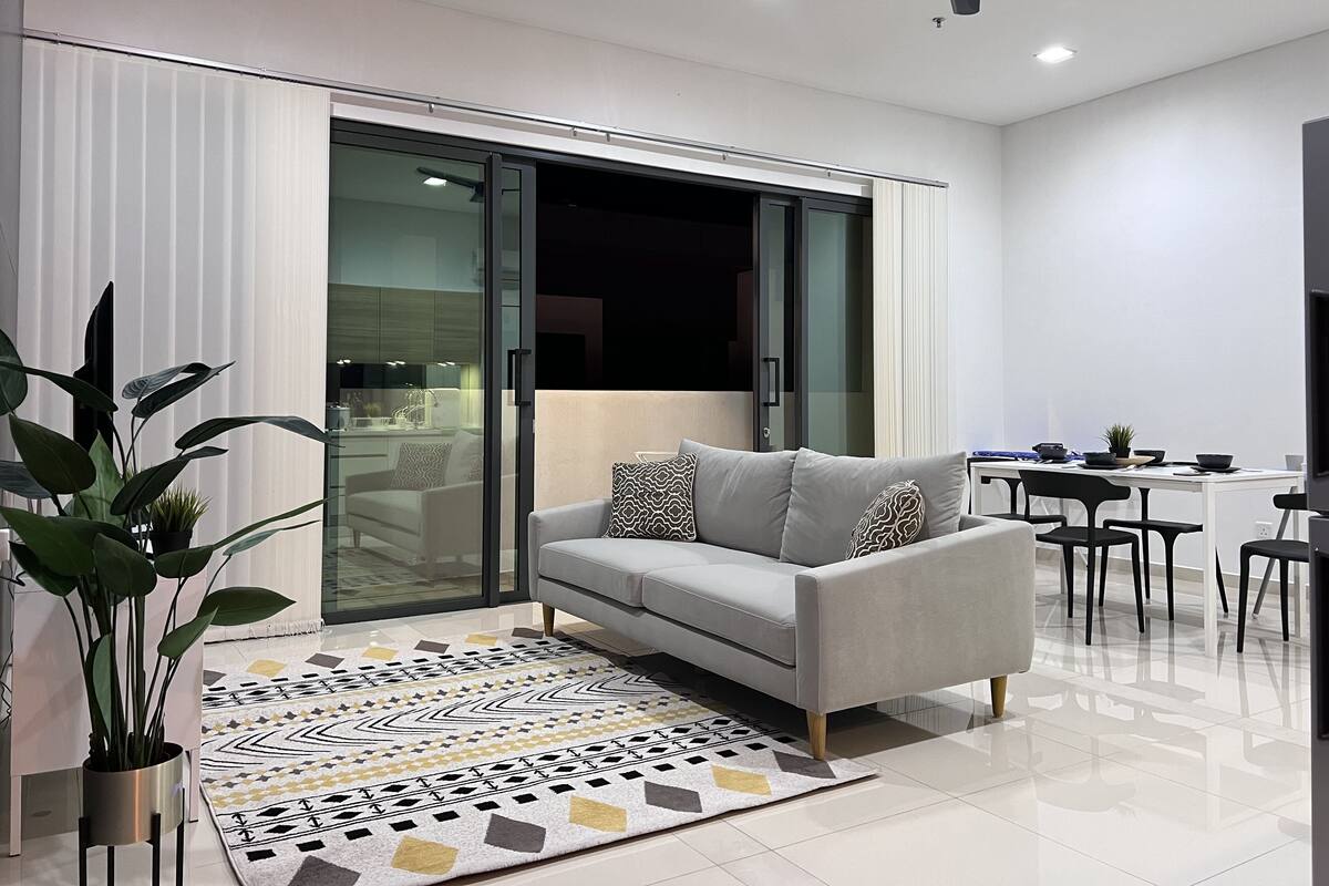 Ein modernes Wohnzimmer mit einem grauen Sofa, einem gemusterten Teppich und einem Essbereich mit Tisch und Stühlen.