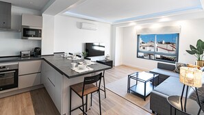 Eine moderne Küche mit einer zentralen Kochinsel, ein Essbereich mit Tisch und Stühlen, eine Sitzecke mit Sofa und ein großes Fenster mit Blick auf Gebäude.