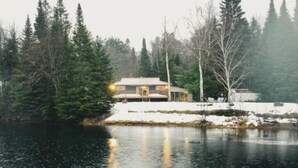 A lakeside cabin with a reflective water surface, surrounded by evergreen trees and a snow-covered ground. - The Mara Waterfront Cottage, Muskoka
 (Huntsville)