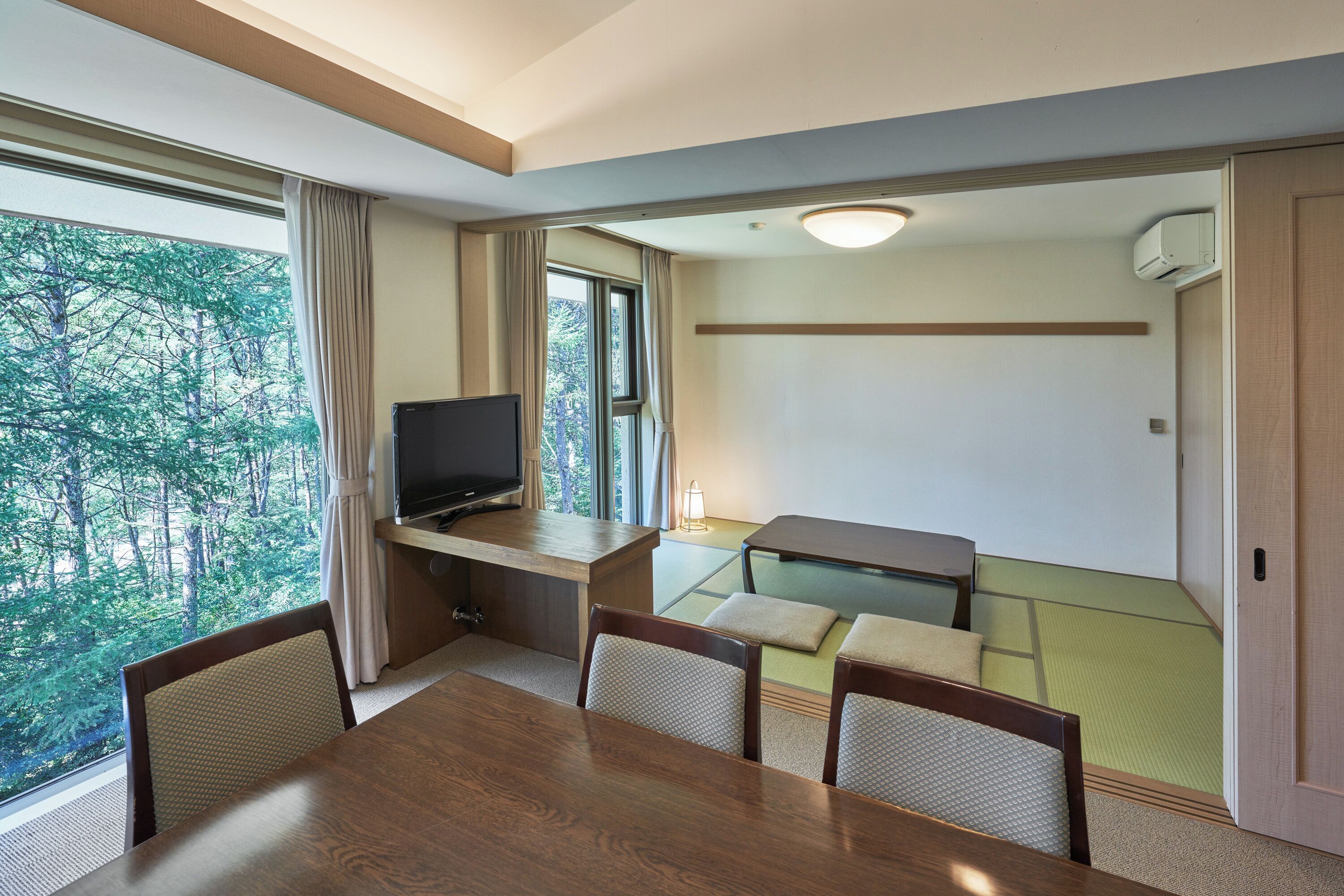 A hotel room with a dining table, chairs, a TV, a sofa, and a view of trees.