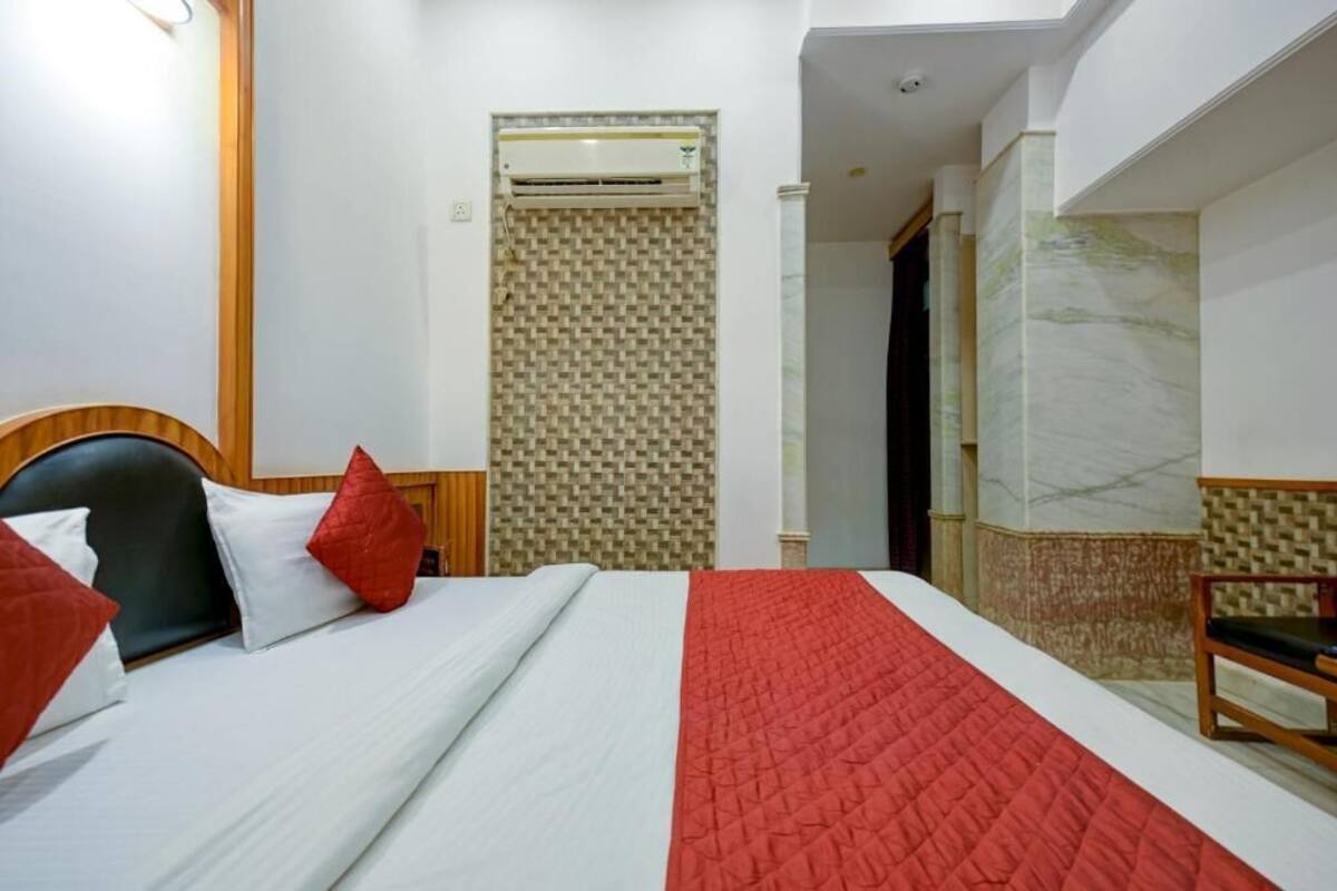 Une chambre d’hôtel avec un lit, une tête de lit en bois, une couverture rouge matelassée, un oreiller rouge et un banc en bois, ainsi qu’un mur orné d’un motif.
