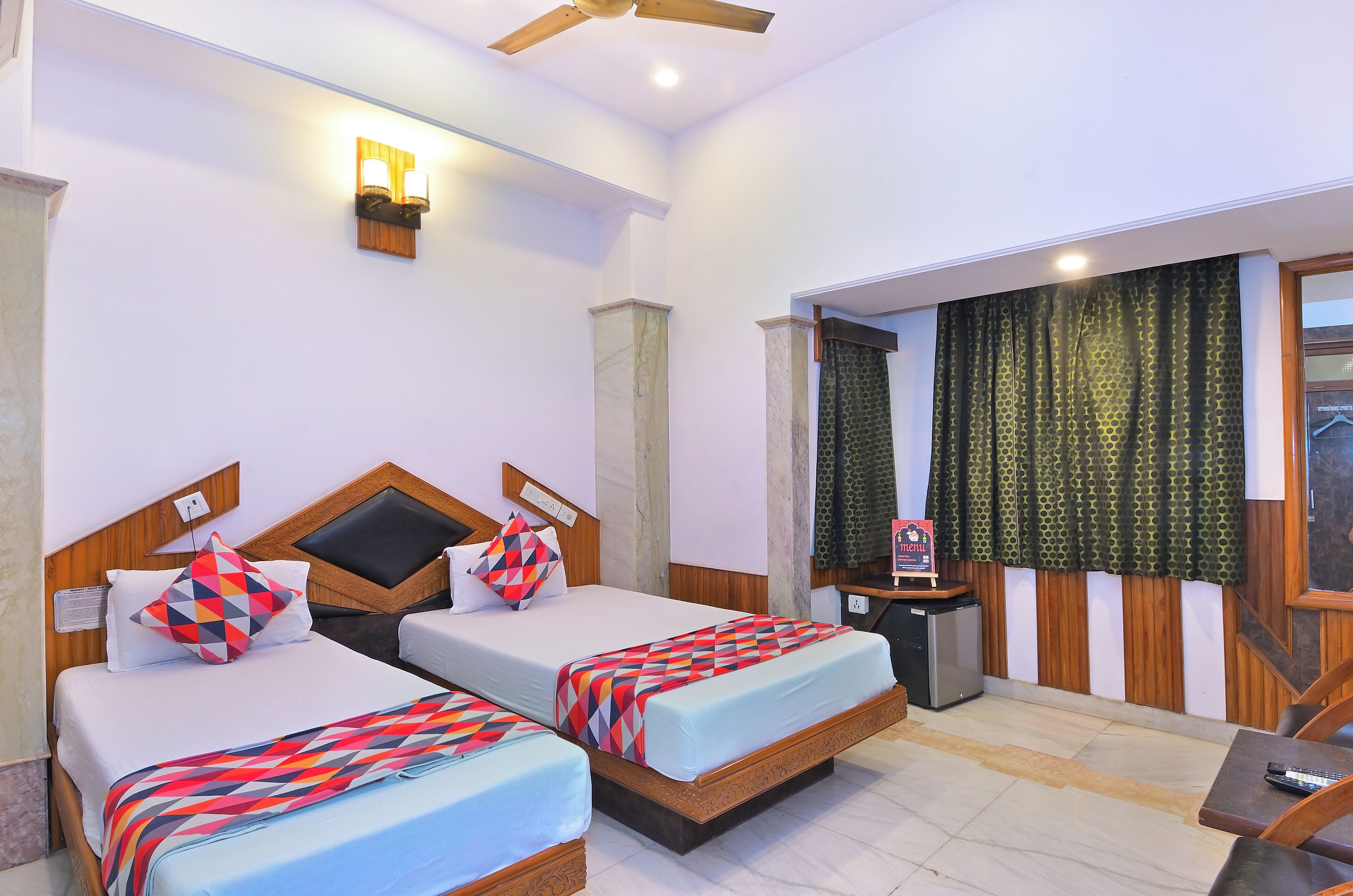A hotel room with two beds, a ceiling fan, a small table, and a window with curtains.