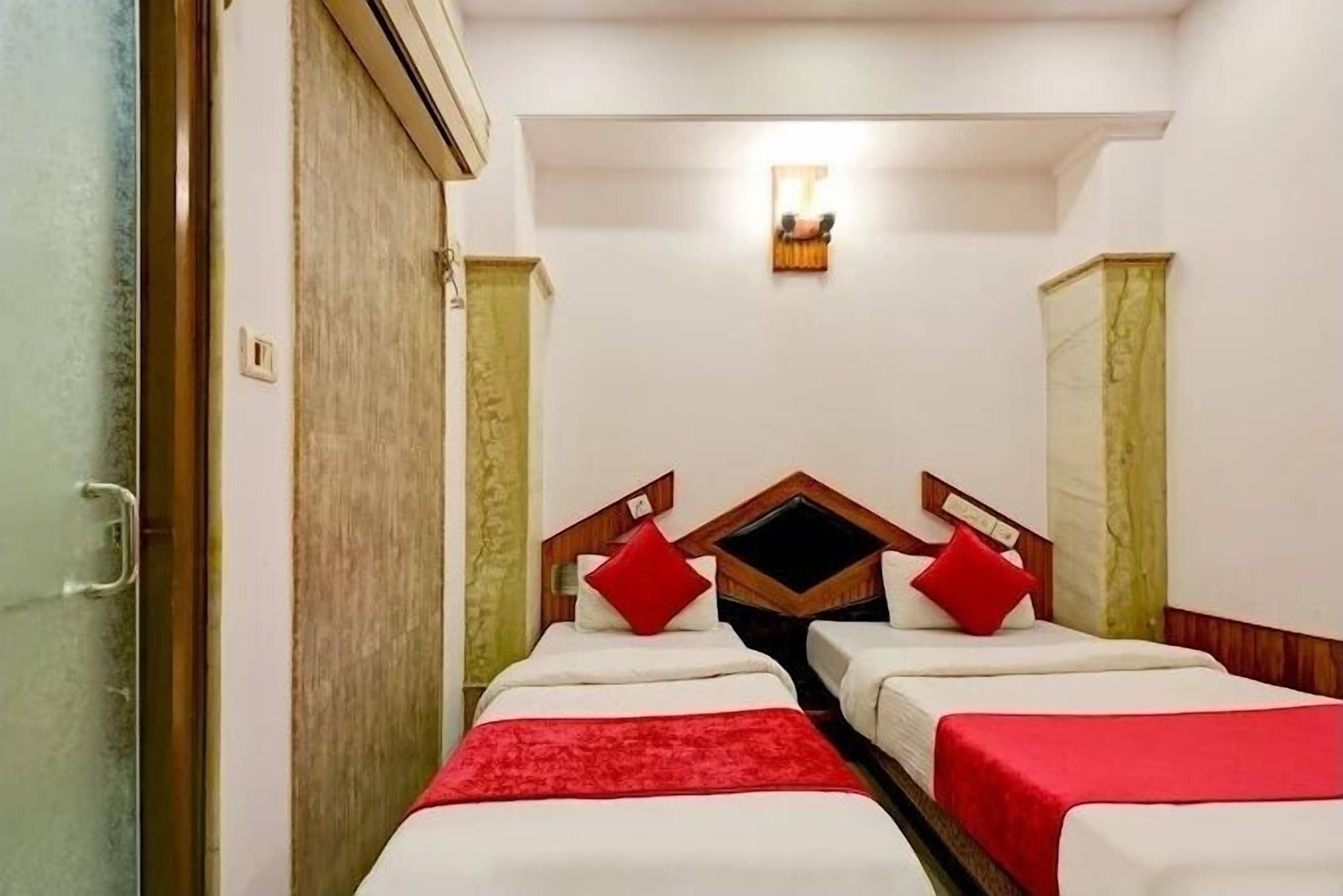 A hotel room with two single beds, each with a red pillow and a white bedspread. There is a small wall-mounted light fixture above the beds.