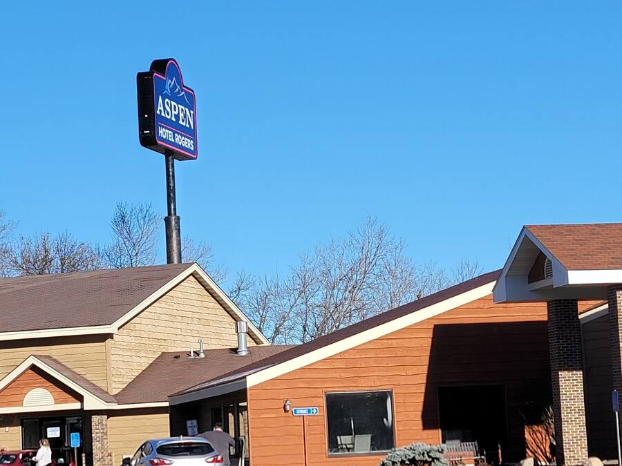 A hotel named 'Aspen Hotel Rogers' with a prominent sign, a parking lot, and a building with a brown roof and beige walls.