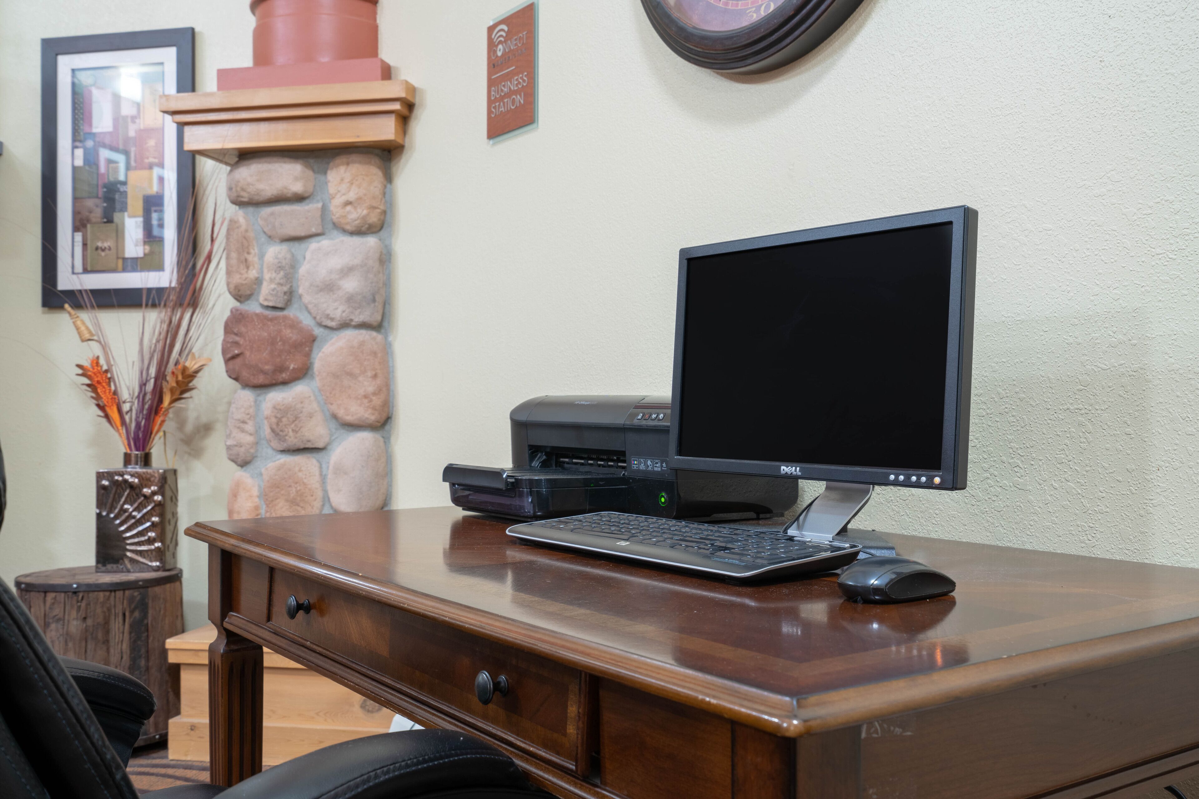 A wooden desk with a computer, keyboard, and mouse. There is a stone wall and a framed picture on the wall.
