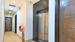 A hallway with a wooden door, a wall-mounted security camera, an elevator, and a vase with flowers. - The Bella Vista – Traveler's Choice Awarded (New Delhi)