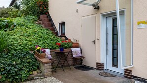 Una casa con fachada blanca, una puerta con panel de vidrio, un pequeño espacio exterior con mesa y sillas, y una escalera que sube a un nivel superior.