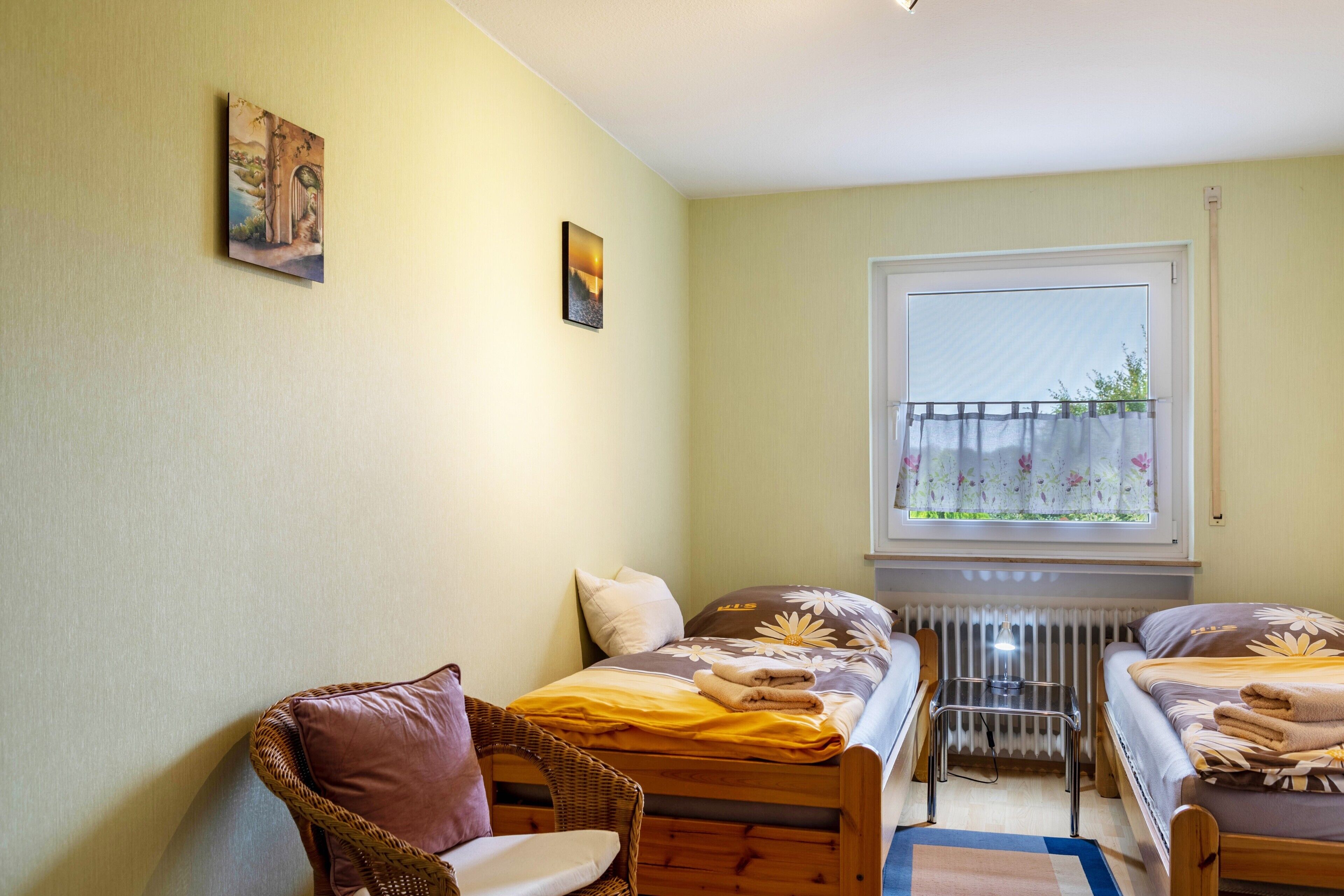 Une petite pièce avec deux lits, une chaise en osier, un radiateur et une fenêtre donnant sur un beau paysage.