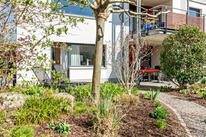 A modern building with a balcony, a garden area with various plants, and a gravel path.