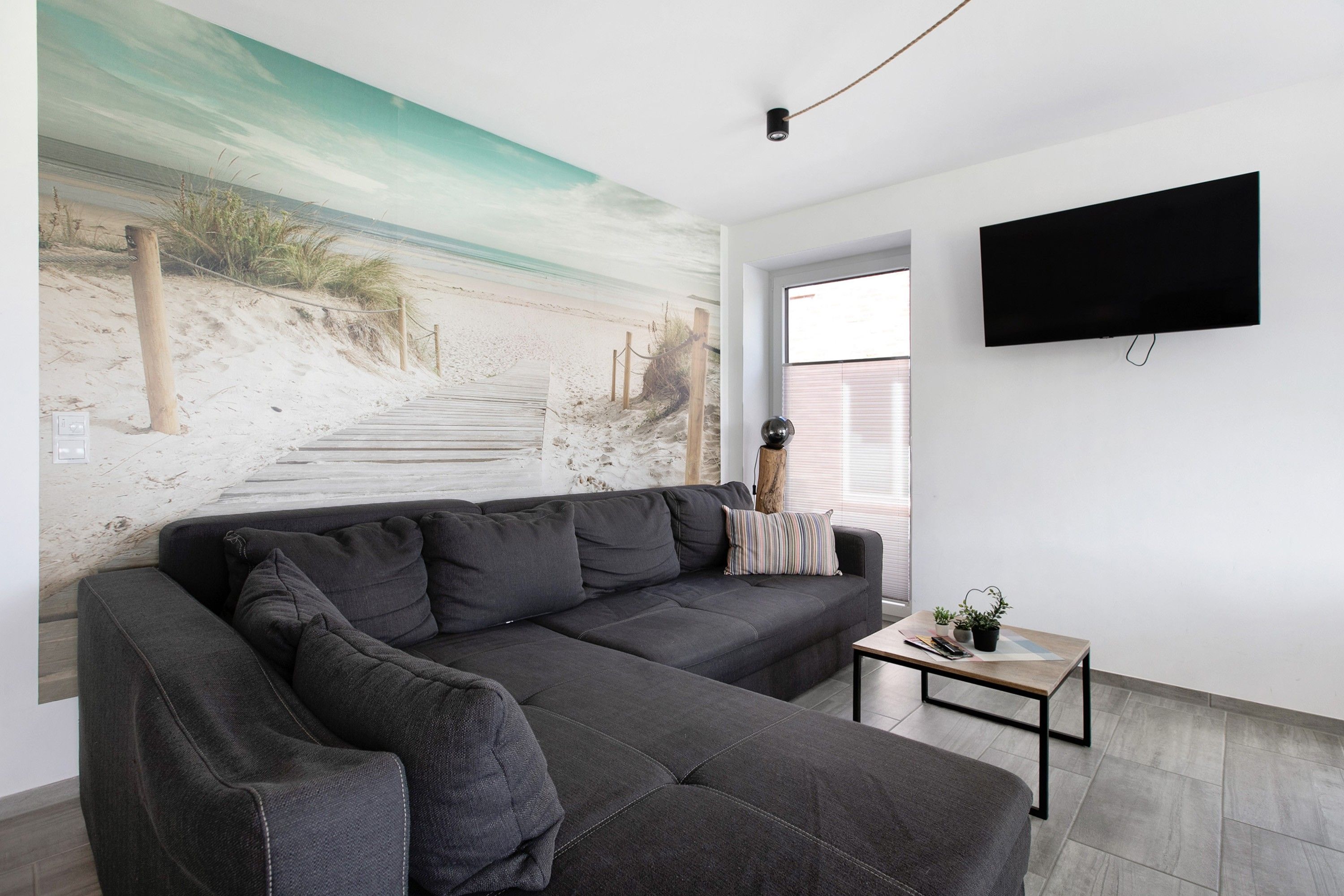 A living room with a large sectional sofa, a small coffee table, a flat-screen TV mounted on the wall, and a beach mural on the wall.