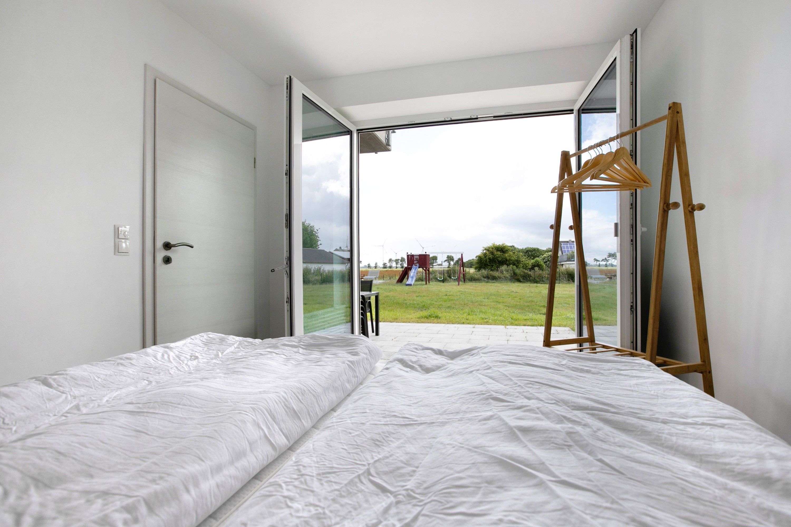 Una camera da letto con un letto, un appendiabiti in legno e una porta-finestra in vetro scorrevole che dà su uno spazio esterno con un'area giochi.