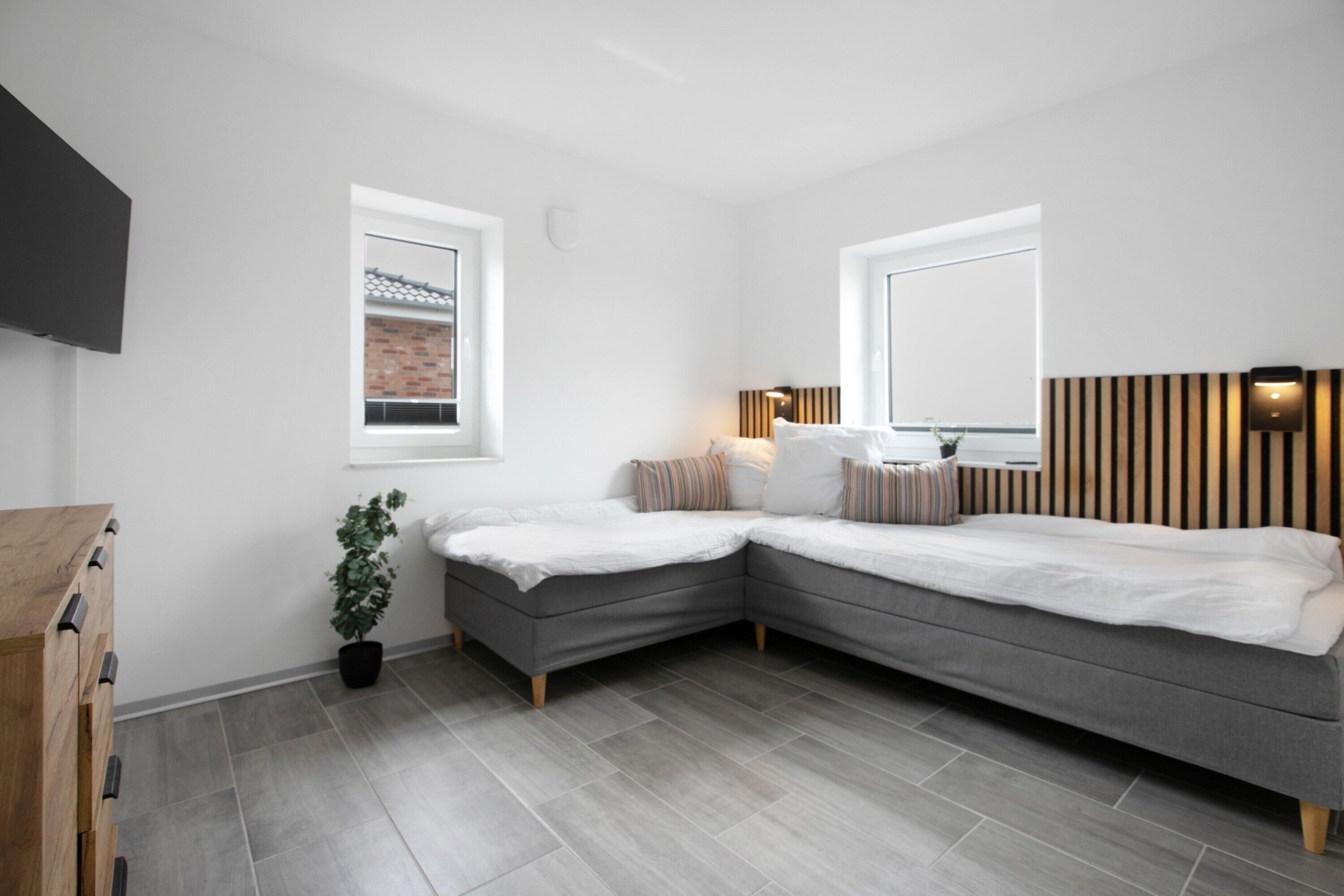 Ein modernes Schlafzimmer mit einem grauen Ecksofa, einem kleinen Nachttisch, einer Topfpflanze und einem an der Wand befestigten Fernseher.