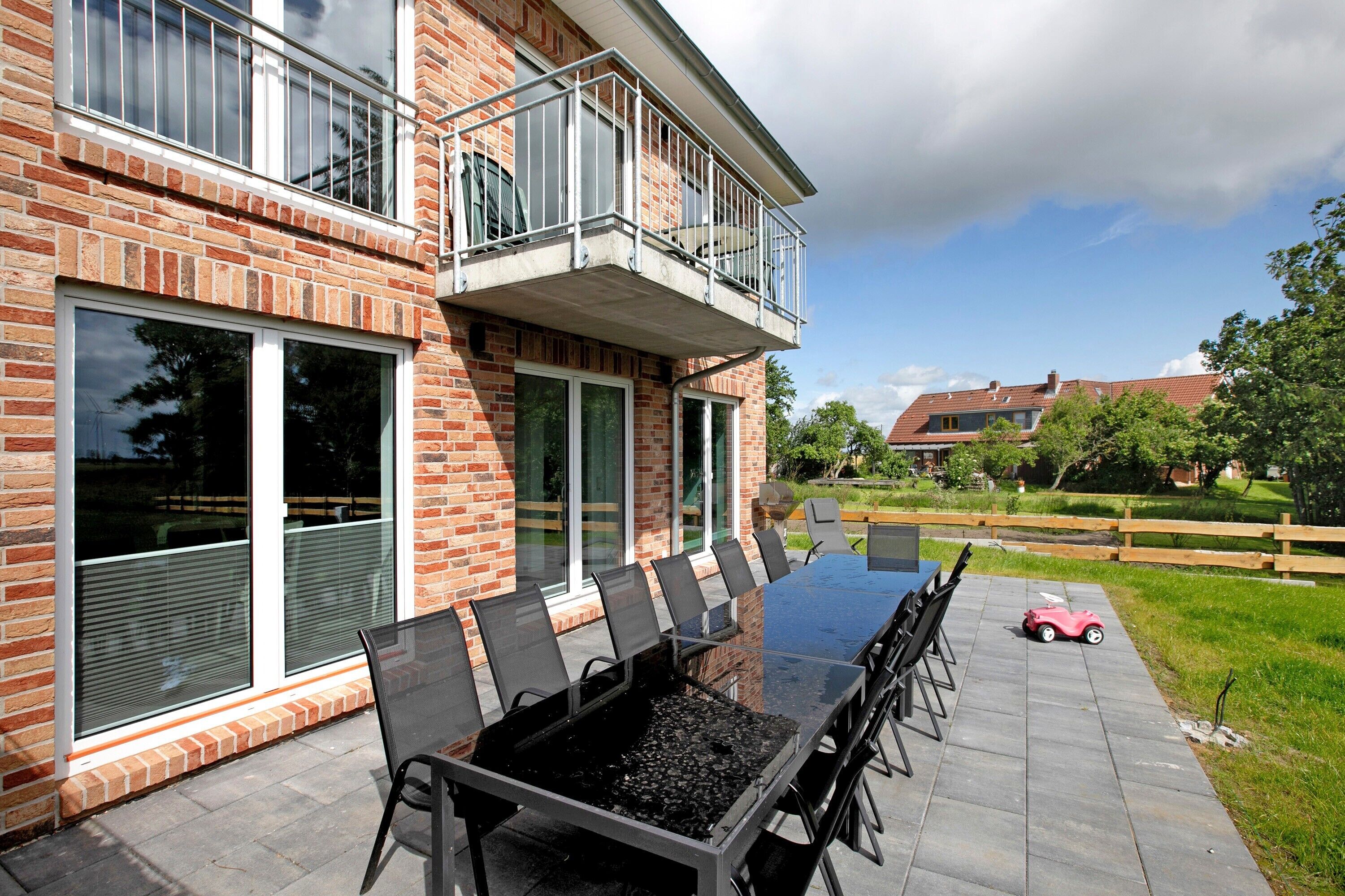 Ein zweistöckiges Backsteinhaus mit Balkon, einer Terrasse mit Tisch und Stühlen mit Glastischplatte und ein kleines rotes Spielzeugauto.