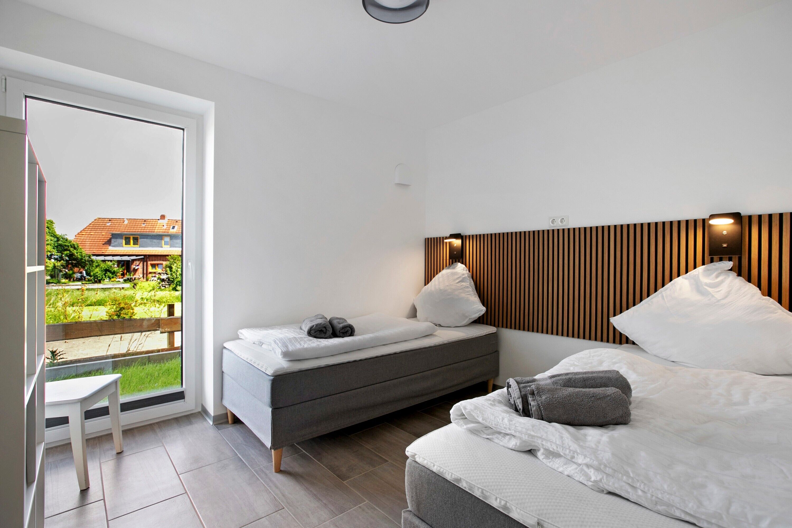 Ein modernes Schlafzimmer mit einem Bett, einer Couch und einem kleinen Tisch. Durch das Fenster ist ein Blick auf einen Garten und Gebäude zu sehen.