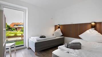 Ein modernes Schlafzimmer mit einem Bett, einer Couch und einem kleinen Tisch. Durch das Fenster ist ein Blick auf einen Garten und Gebäude zu sehen.