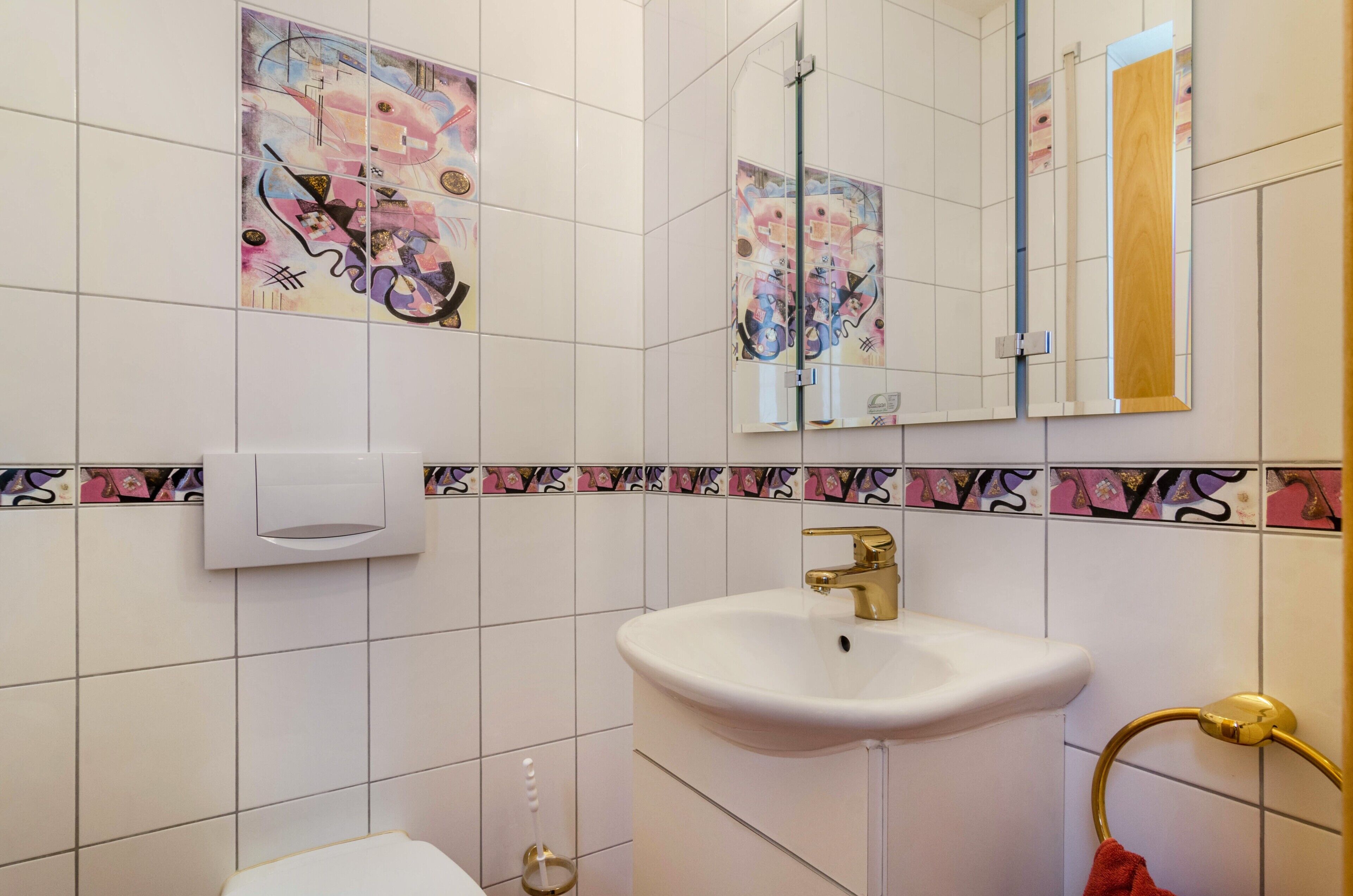 Kylpyhuone, jossa on valkoinen pesuallas, kultainen hana, wc-istuin ja peili.
