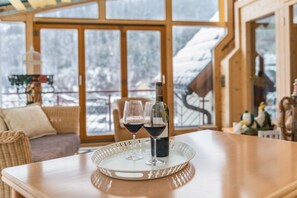 A wooden table with a tray holding two wine glasses and a bottle, set in a room with large windows and wicker furniture.
