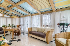 Ein Zimmer mit einer Couch, Esstisch, Stühlen, einer Decke mit Oberlichtern und Vorhängen.
