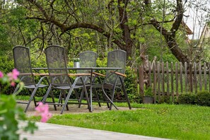 Um pátio de jardim com quatro cadeiras de metal e uma mesa, cercado por vegetação e um muro de madeira.