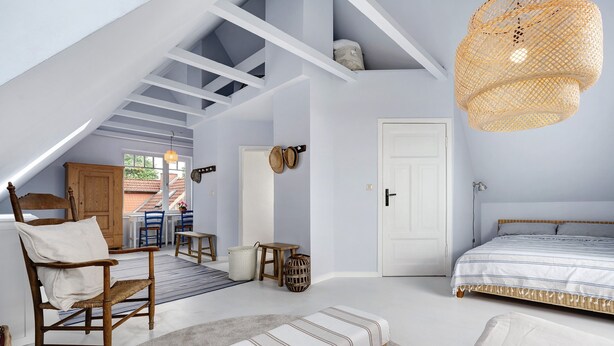 Quarto em estilo loft com teto inclinado, cama com roupa de cama listrada, cadeira de madeira, um banco e luminária pendente de vime.