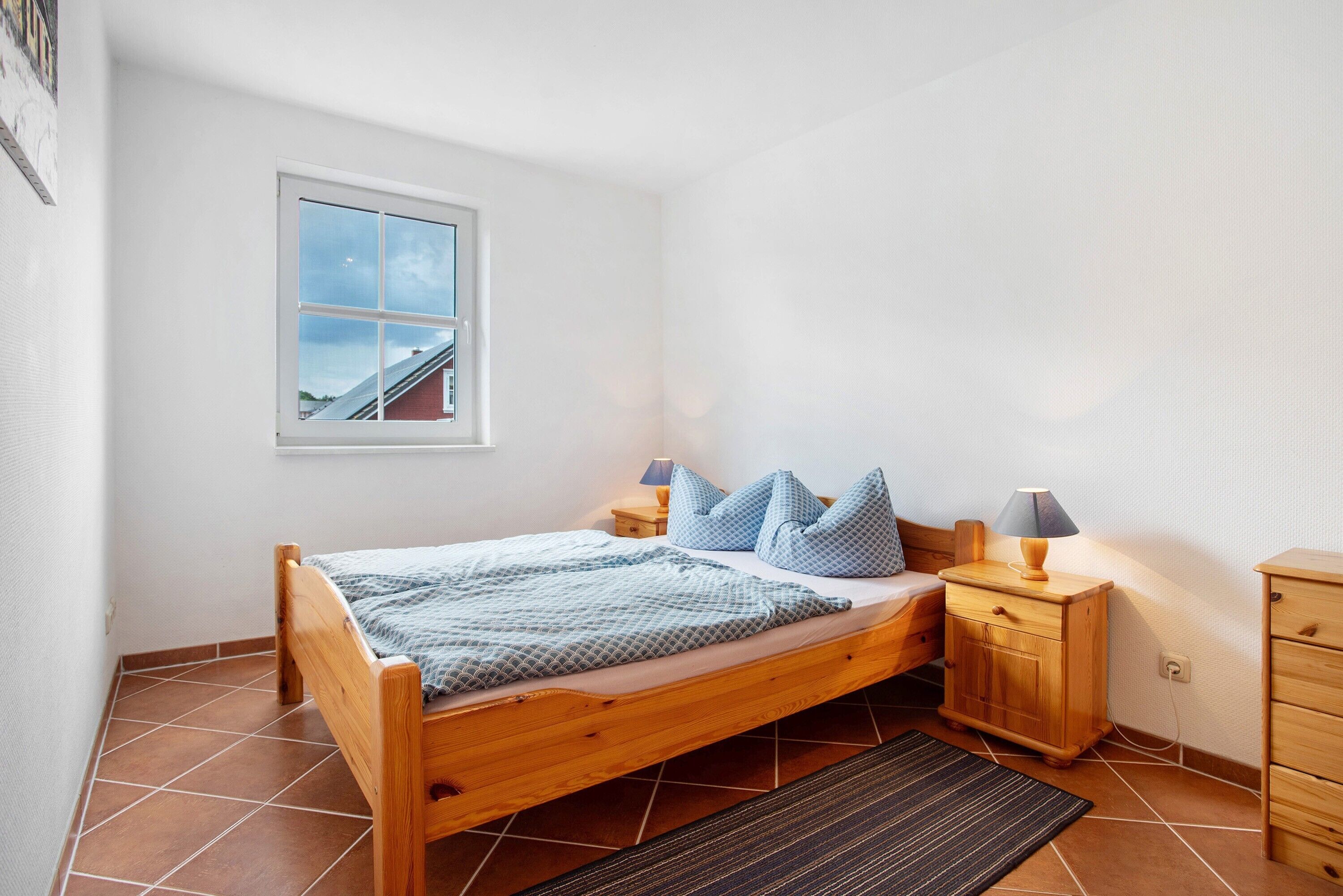 A bedroom with a wooden bed, a nightstand, a dresser, a window with a view, and a tiled floor.