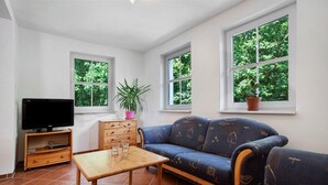 Ein Wohnzimmer mit einer Couch, einem Fernseher, einem Tisch und Fenstern mit Blick auf Bäume.