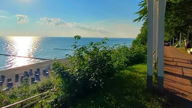 A beach with sunlit water, a wooden walkway, and greenery.