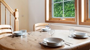 A dining table set with plates, bowls, glasses, and cutlery, a window with a view of greenery, and wooden chairs.