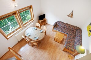 Une pièce comprenant une table de salle à manger ronde en bois, deux chaises en bois, un canapé, un téléviseur posé sur un meuble en bois et une fenêtre donnant sur un paysage verdoyant.