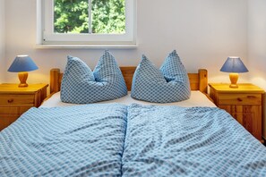 Un lit recouvert d’une couvre-lit bleue à motifs, deux oreillers assortis, des tables de chevet en bois avec des lampes, et une fenêtre donnant sur un paysage verdoyant.