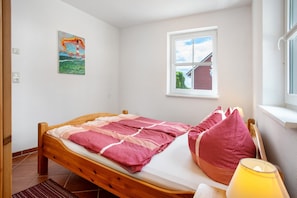 A single bed with a wooden frame, a window with a view of a house, and a painting on the wall.