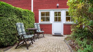 Un bâtiment rouge avec une porte et des fenêtres blanches, une terrasse pavée avec deux chaises et une haie.