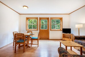 Una habitación con mesa de comedor, sillas, sofá, televisión y ventanas con vistas a zonas verdes.