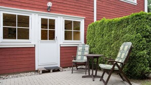 Una casa de madera roja con puerta y ventanas blancas, un patio empedrado, dos sillas y una pequeña mesa.