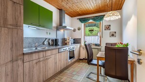 A compact kitchen with wooden cabinets, a stove, a microwave, and a dining area with a chair and table. - Apartment 'Ferienwohnung Rehsiepen' with Mountain View, Private Terrace and Wi-Fi (Schmallenberg)