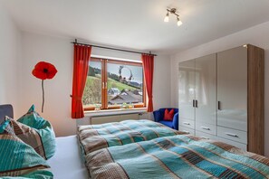 A bedroom with a large bed, a wardrobe, a window with a view of a house, and a red flower on the wall.