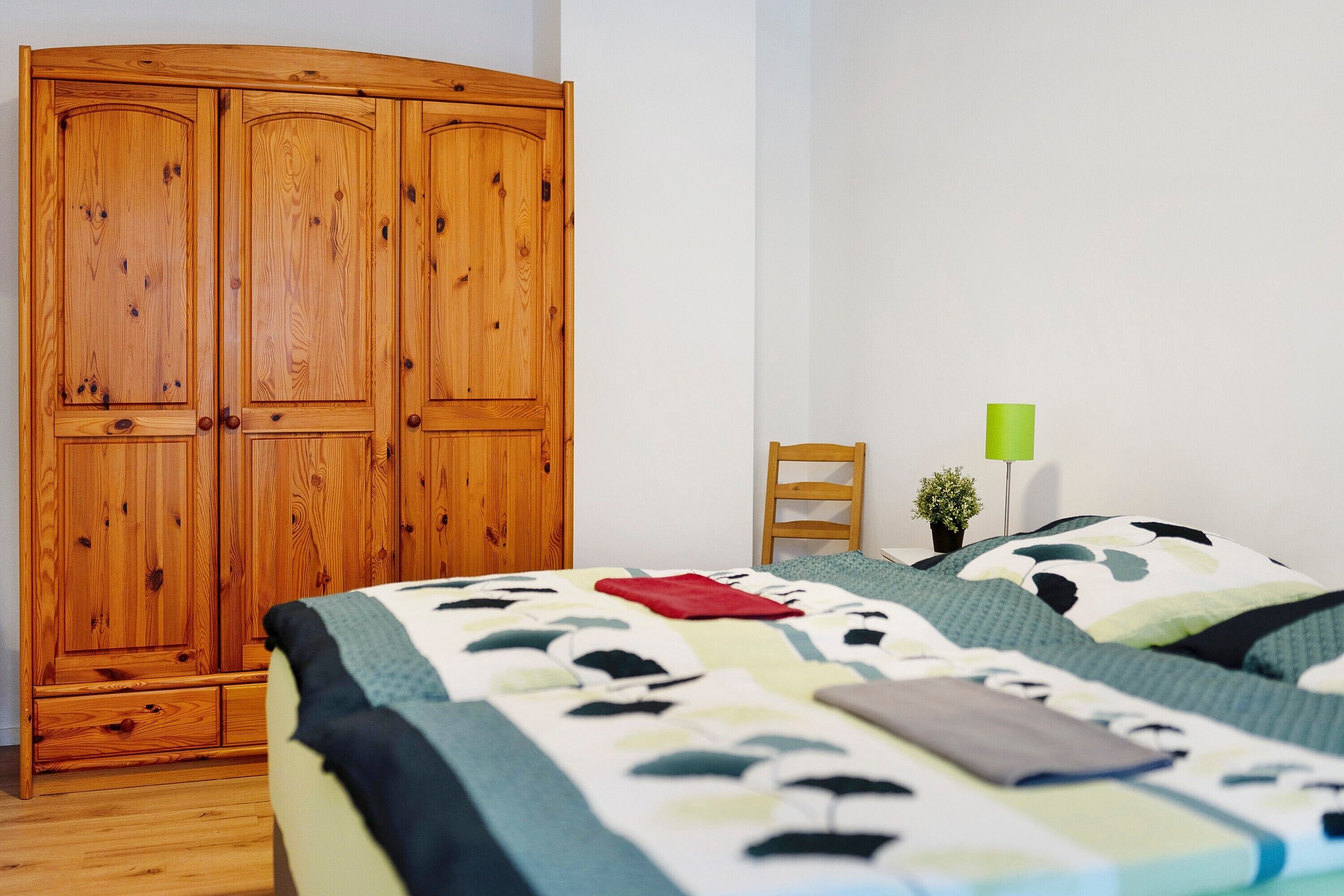 Een slaapkamer met een grote houten garderobe, een bed met een bedovertrek met dessin, een klein nachtkastje met een lamp en een stoel.