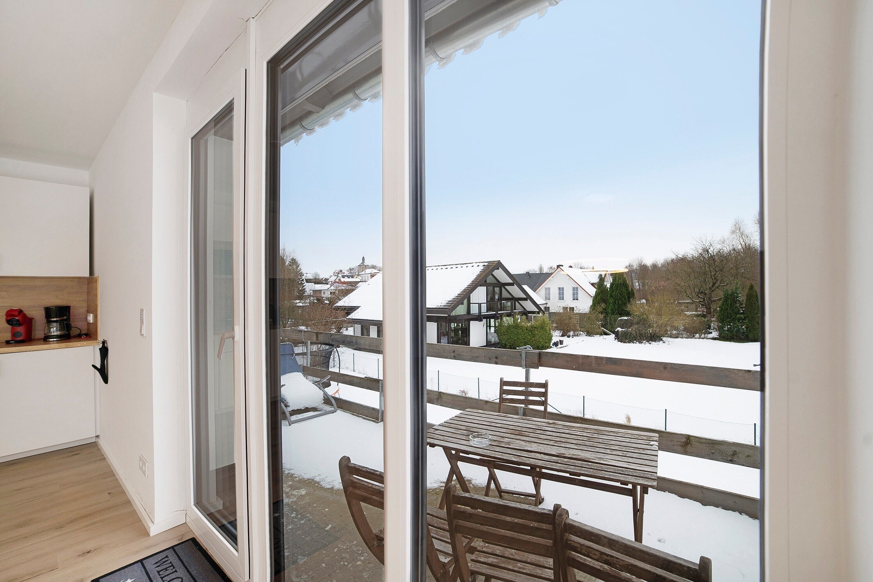 A balcony with a wooden table and chairs, a view of a snowy landscape, and a house in the background.
