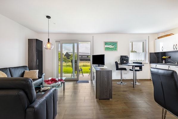 A modern living room with a sofa, a glass coffee table, a TV, a desk with a computer, and a kitchen area with a counter and chairs.