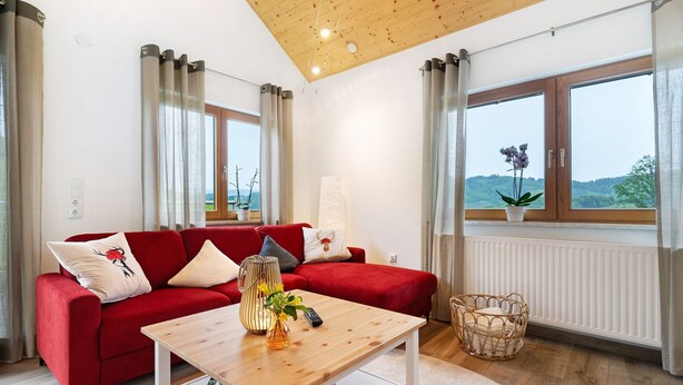 Un sofá rojo con cojines, una mesa de centro de madera, una cesta de mimbre y vistas a zonas verdes a través de amplios ventanales.