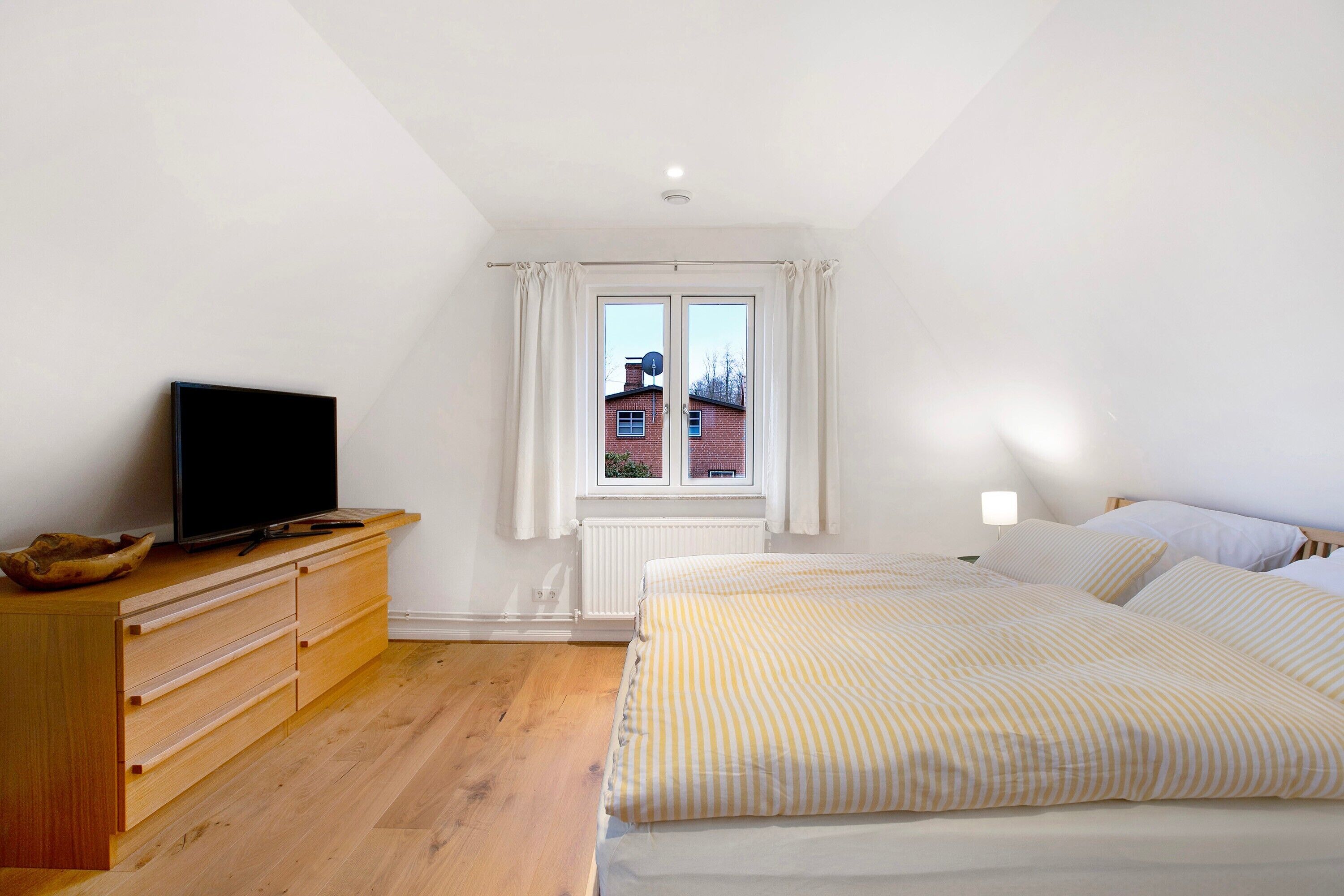 Een slaapkamer met een bed, een houten dressoir, een televisie, een raam met gordijnen en een radiator.