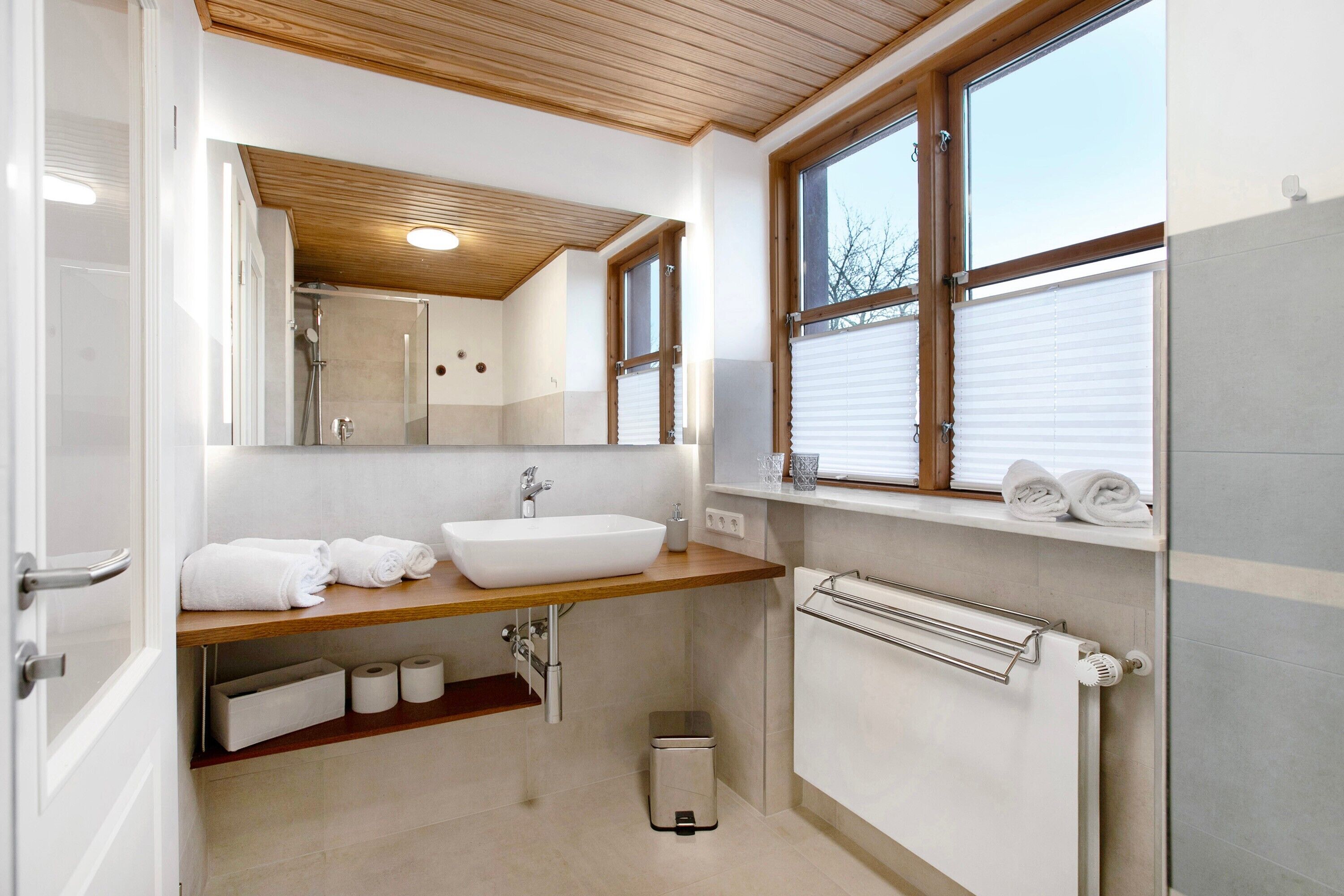 A modern bathroom with a wooden vanity, a large mirror, a white sink, and a window with blinds.