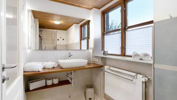 A modern bathroom with a wooden vanity, a large mirror, a white sink, and a window with blinds.